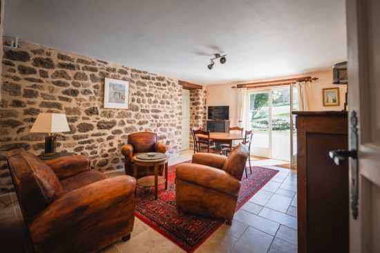 Living room and dining room area of ​​the L’Heure Exquise lodge, featuring leather armchairs, a red carpet, natural stone walls and a large glass door opening onto the garden.
