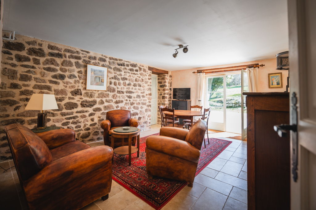 Living room and dining room area of ​​the L’Heure Exquise lodge, featuring leather armchairs, a red carpet, natural stone walls and a large glass door opening onto the garden.