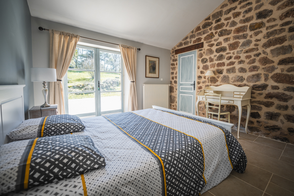 Interior of the Aurore room at the L’Heure Exquise lodge with a double bed, an exposed stone wall, and a large bay window offering a view of the green garden.