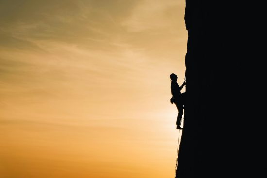 Photo d’un escaladeur grimpant une falaise, silhouette visible à contre-jour sous un ciel lumineux.