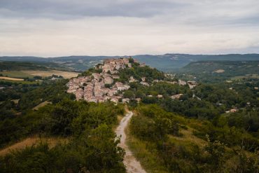 Photo of the medieval village of Cordes-sur-Ciel seen from afar, perched on a hill surrounded by greenery.