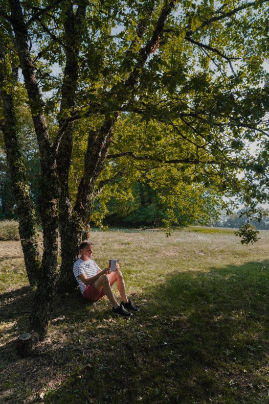 Photo de Thierry, l’hôte du gîte L’Heure Exquise, assis par terre et adossé à un arbre, lisant un livre dans le jardin